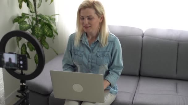 Χαμογελαστή γυναίκα που ηχογραφεί το βίντεο μπλογκ της. Μια ενήλικη γυναίκα μιλάει μπροστά σε μια βιντεοκάμερα για το κανάλι της στο blog. Γυναίκα μπλόγκερ βιντεοσκοπεί το βίντεο στο σπίτι της. Vlogger κάνει online streaming χρησιμοποιώντας smartphone. — Αρχείο Βίντεο