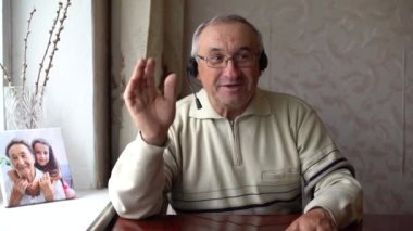 Gülümseyen son sınıf öğrencisi kulaklık takıyor, kameraya kulaklık takıyor dizüstü bilgisayarda video görüşmesi yapıyor, kulaklık takan mutlu yaşlı erkek evde oturup modern teknolojileri ve kablosuz bağlantıyı kullanıyor.