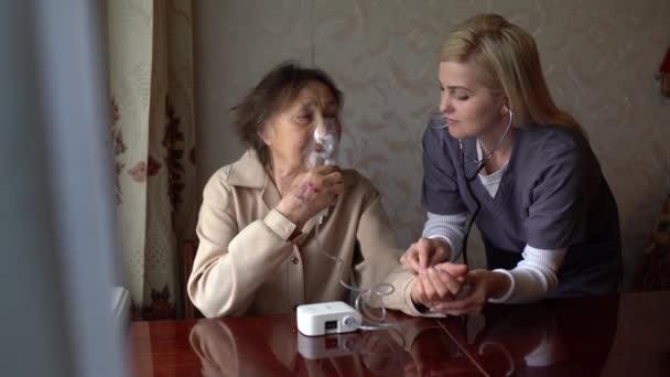 老女患者の顔にネブライザーマスクをつけて自宅で吸入する若い熟練した快適な女性医師。インフルエンザ、風邪、咳の治療. — ストック動画