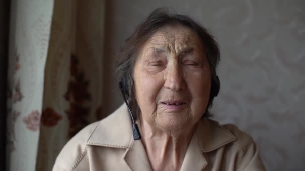 Очень старая женщина в наушниках ведет видеоразговор у себя дома. Концепция видеоразговора. headshot online. — стоковое видео