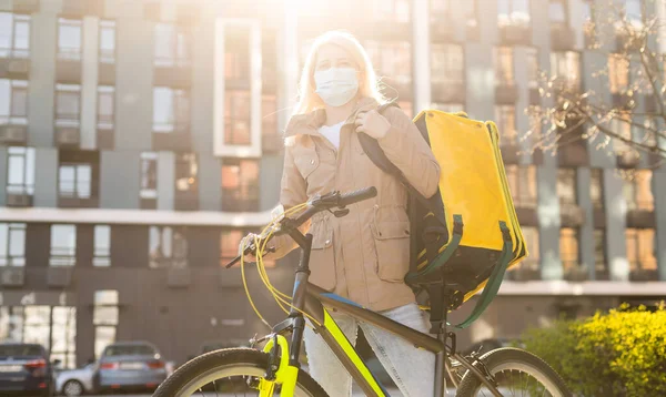 女信使戴着医疗面罩，背着黄色的热背包，骑着自行车在城市里运送食物。食品交付服务概念. — 图库照片