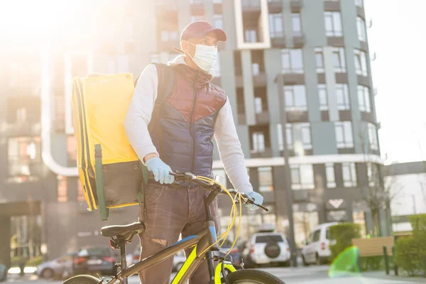 年轻的信使戴着医疗面罩，背着黄色的热背包，骑着自行车在城市里运送食物。食品交付服务概念 — 图库照片