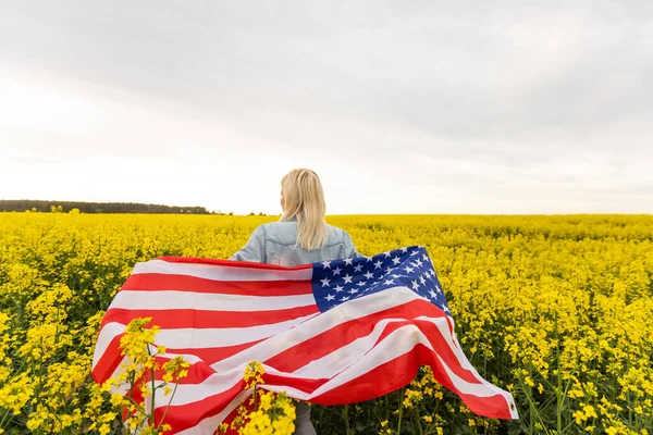 Mujer adulta sosteniendo bandera americana con poste, estrellas y rayas en un campo de colza amarillo. Bandera de EE.UU. ondeando en el viento — Foto de Stock