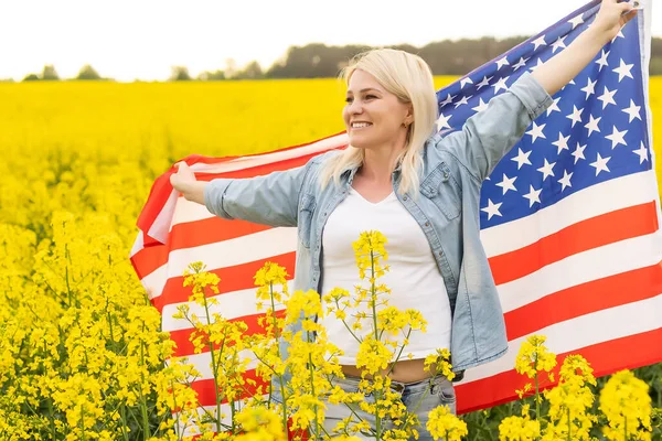 Mujer adulta sosteniendo bandera americana con poste, estrellas y rayas en un campo de colza amarillo. Bandera de EE.UU. ondeando en el viento — Foto de Stock