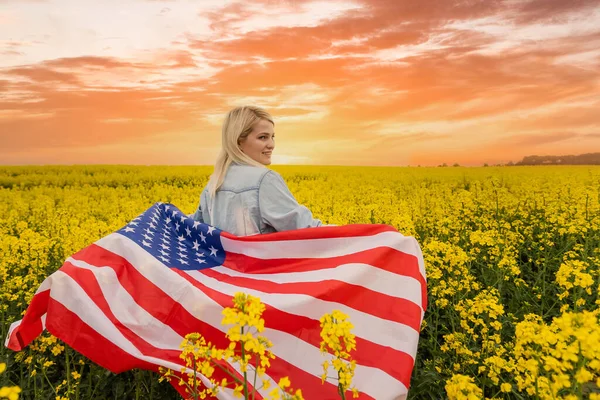 바람에 옷자락에 미국 국기를 들고 있는 매력적 인 여성. 푸른 하늘을 배경으로 여름 풍경. 수평 방향. — 스톡 사진