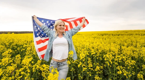 Kolza tohumu tarlasında rüzgarda Amerikan bayrağı tutan çekici bir kadın. Mavi gökyüzüne karşı yaz manzarası. Yatay yönelim. — Stok fotoğraf
