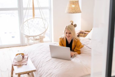 Uzaktan kumandalı ev işi, karantina sırasında serbest çalışma. Gülümseyen bayan rahat oturma odasında dizüstü bilgisayarda oturuyor ve çalışıyor.