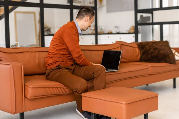 Giovane uomo sorridente, legge lo schermo di un computer portatile mentre si rilassa su un comodo divano a casa — Foto Stock