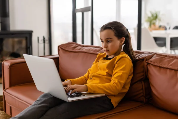 Smart Little Girl hace la tarea en su sala de estar. Ella se sienta y utiliza el ordenador portátil. — Foto de Stock