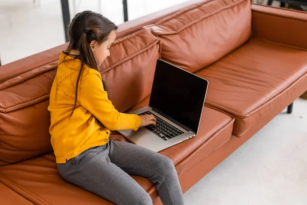 Slimme kleine meid doet huiswerk in haar woonkamer. Ze zit en gebruikt laptop. — Stockfoto