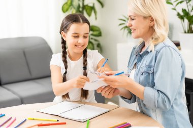 Anne ve kız güzel ve mutlu anneler günü için kağıt ve kalemlerle birlikte masaya oturarak bir kart yapın.