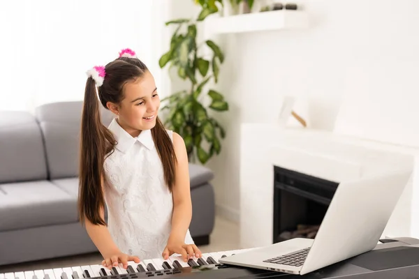 어린 소녀가 컴퓨터를 통해 학교 선생님에 의해 인터넷 음악 교실에 연결되는 피아노를 배우고 있습니다. 새로운 생활 방식 과 교육, 가정에서 공부하는 학생. — 스톡 사진