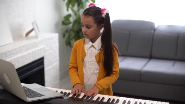 Een klein meisje dat piano speelt. Online muziekles met leraar via laptop — Stockvideo