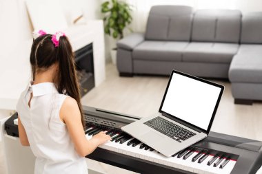 Evde eğitim gören küçük bir kız, okul öğretmeninin internet müziğine bağlı bilgisayardan piyano öğreniyor. Yeni normal yaşam tarzı ve eğitim, ev konseptinde öğrenim.