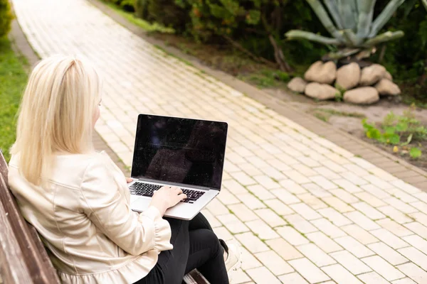 Jonge aantrekkelijke zakenvrouw met behulp van een pc laptop computer tijdens het zitten op een houten bank in een stadspark. — Stockfoto