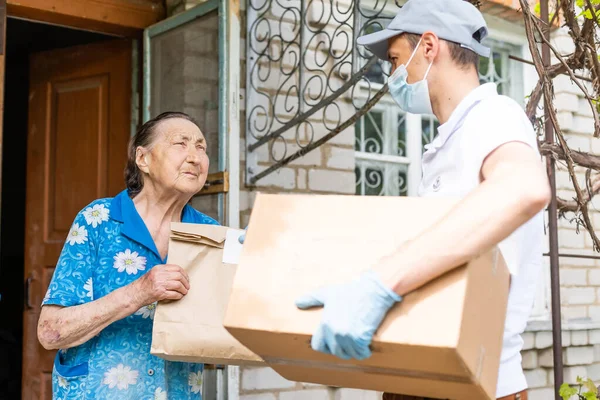 Joven voluntario con máscara le da a una anciana cajas con comida cerca de su casa. El hijo ayuda a una madre anciana soltera. Apoyo familiar, cariño. Cuarentena, aislada. Coronavirus covid-19 —  Fotos de Stock