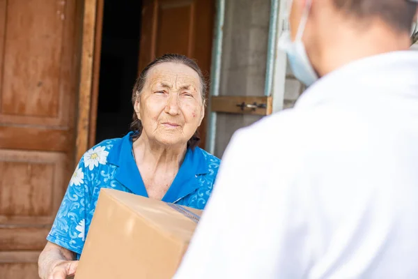 Young male volunteer in mask gives an elderly woman boxes with food near her house. The son helps a single elderly mother. Family support, caring. Quarantined, isolated. Coronavirus covid-19 — Stock Photo, Image
