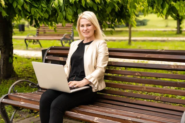 Junge attraktive Geschäftsfrau sitzt mit einem PC-Laptop auf einer Holzbank im Stadtpark. — Stockfoto