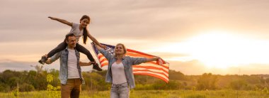 Kızlarıyla birlikte gün batımında kırsalda Amerikan bayrağı taşıyan genç ebeveynler. Bağımsızlık Günü kutlamaları