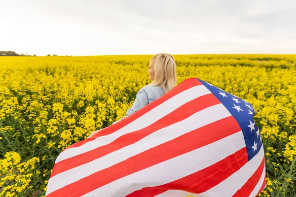 Vuxen kvinna som håller amerikansk flagga med stång, stjärnor och rand i ett gult rapsfält. USA flagga fladdrar i vinden — Stockfoto