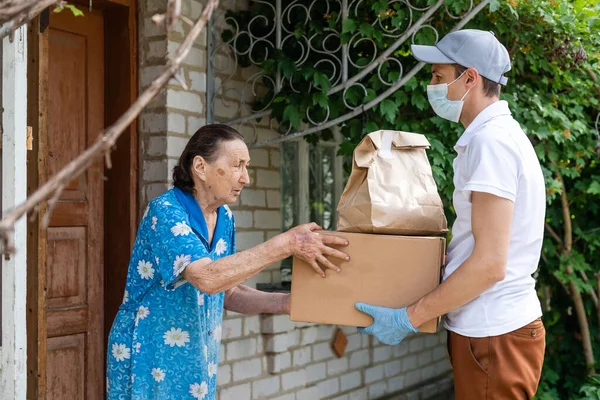 Young male volunteer in mask gives an elderly woman boxes with food near her house. The son helps a single elderly mother. Family support, caring. Quarantined, isolated. Coronavirus covid-19