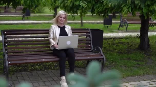 Vit kvinna i headset som skriver på datorn på bänken i parken. Kvinnlig student gör konferens videosamtal på bärbar dator samtal med webb handledare, online lärare i webcam chat. — Stockvideo