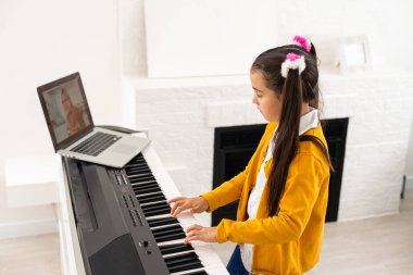 Küçük kız evde dizüstü bilgisayarla sınıf eğitiminde piyano çalıyor. Covid-19 kriz karantinasında öğreniyor.
