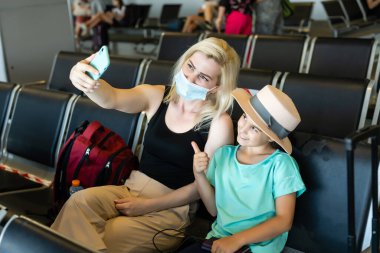 Anne ve kız turist hijyen maskesi takıyor ve havaalanı terminalinde oturuyor. Yeni normal yolcuların güvenliği ve sosyal mesafeler için fikir.