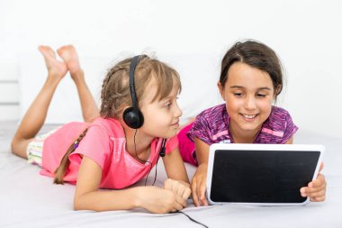 İki küçük tatlı kız dijital tablet kullanıyor ve çocuk odasında yatakta yatarken gülümsüyor.