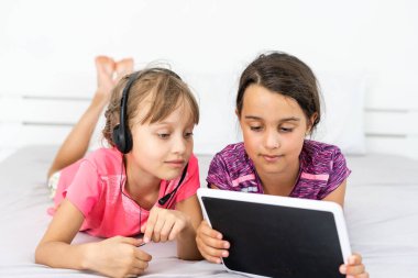 İki küçük tatlı kız dijital tablet kullanıyor ve çocuk odasında yatakta yatarken gülümsüyor.