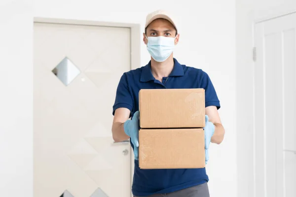 Man from delivery service in t-shirt, in protective mask and gloves giving food order and holding boxes over white background.