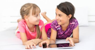 İki küçük tatlı kız dijital tablet kullanıyor ve çocuk odasında yatakta yatarken gülümsüyor.