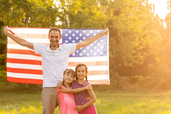 Familia feliz sentados juntos en su patio trasero sosteniendo la bandera americana detrás de ellos. Pareja sonriente con sus hijos celebrando el Día de la Independencia Americana con bandera americana — Foto de Stock