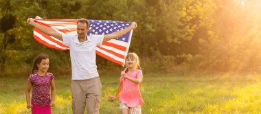 Arka bahçelerinde birlikte oturan mutlu aile arkalarında Amerikan bayrağını taşıyor. Amerika 'nın bağımsızlık gününü çocuklarıyla kutlayan gülümseyen çift Amerikan bayrağı taşıyor.