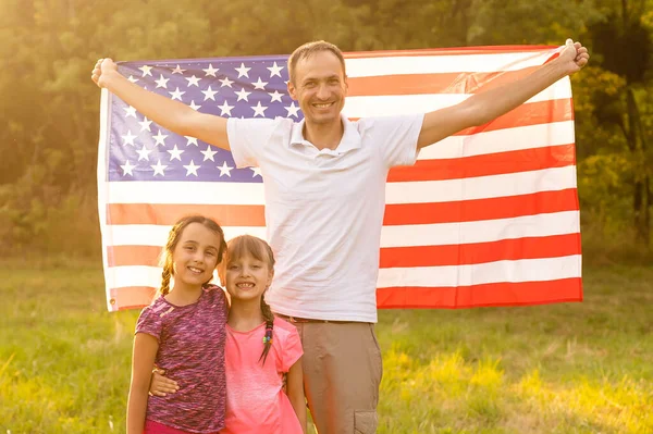 Familia feliz sentados juntos en su patio trasero sosteniendo la bandera americana detrás de ellos. Pareja sonriente con sus hijos celebrando el Día de la Independencia Americana con bandera americana — Foto de Stock