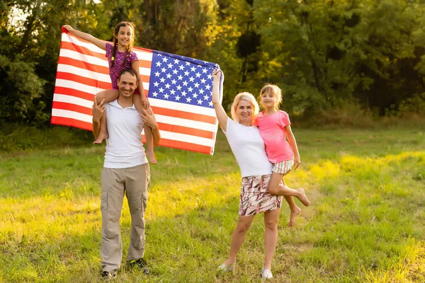 Familia feliz con la bandera americana en un campo de trigo al atardecer. Día de la Independencia, 4 de julio. — Foto de Stock