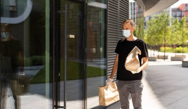 COVID-19 pandemik tecrit konsepti sırasında temassız teslimat. Maske ve eldiven takan kurye bir paket kutusu tutuyor ve akıllı telefondan adres bilgilerini kontrol ediyor.