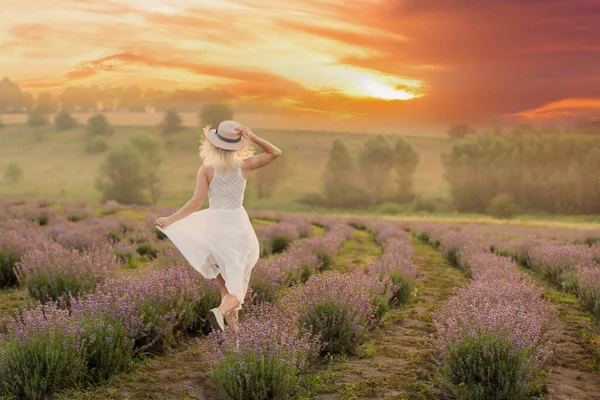 ラベンダー畑を楽しそうに駆け抜ける白いドレスを着た美しい若い健康な女性、麦藁帽子 — ストック写真