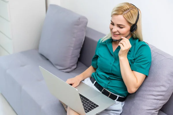 Uśmiechnięta młoda kobieta ze słuchawkami i laptopem na sofie. Pracuje w domu w zamknięciu kwarantanny. Samoizolacja na dystans społeczny — Zdjęcie stockowe