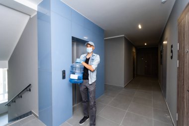 Yüz maskeli bir kurye. Girişte bir şişe soğuk su ve pano var. Koronavirüs karantinası sırasında teslimat