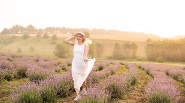 Beyaz elbiseli, genç ve güzel bir kadın lavanta tarlasında neşeyle koşuyor. Hasır şapkalı.