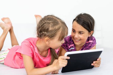 İki küçük tatlı kız dijital tablet kullanıyor ve çocuk odasında yatakta yatarken gülümsüyor.