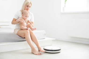 Genç anne yeni yürümeye başlayan oğluyla kanepede oturuyor ve robot elektrik süpürgesinin ev işi yapmasını izliyor..