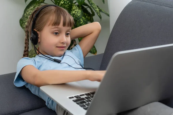 Дистанционное обучение. Веселая маленькая девочка в наушниках с помощью ноутбука изучает через систему электронного обучения онлайн. — стоковое фото