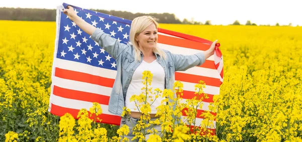 Mujer atractiva sosteniendo una bandera americana en el viento en un campo de colza. Paisaje de verano contra el cielo azul. Orientación horizontal. — Foto de Stock