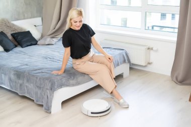Yerleri temizlemek için otomatik süpürge kullanan genç bir kadın makine ev işi robotu kontrol ediyor.