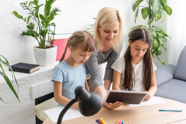 Anne ve iki kız evde ödev yapıyor ve kitap okuyor.