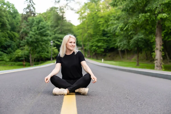 Joyful jovem senhora em roupas casuais andando ao longo da estrada de asfalto no campo, carona para passeio ao ar livre. Adorável mulher milenar viajando sozinha por autostop — Fotografia de Stock