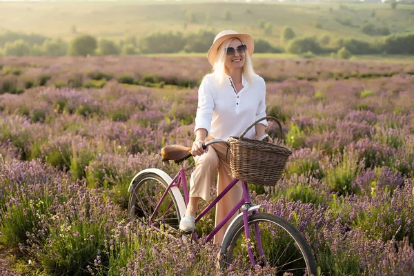 Mulher saudável jovem bonita com um vestido branco correndo alegremente através de um campo de lavanda segurando um chapéu de palha sob os raios do sol poente — Fotografia de Stock