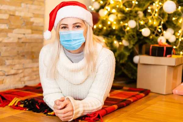 Coronawirus salgınıyla Noel. Koruyucu yüz maskesi takan ve arka planda Noel ağacı, koronavirüs ve Noel konsepti olan Covid-19 için üzgün görünen genç bir kadının portresi. — Stok fotoğraf